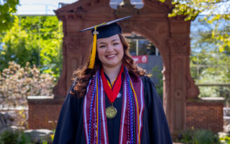 A woman in a graduation cap and gown stands in front of a brick arch outdoors.