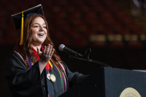 A woman in a black cap and gown stands at a microphone and speaks. She is smiling.