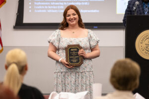 A woman stands, holding a plaque, and smiles. She is inside. Two people face her, sitting down.