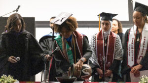 A woman in a graduation cap and gown pours water from a vase into a bowl on a table inside. Two other graduates watch, as a woman to the left holds a microphone.