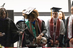 A woman in a graduation cap and gown pours water from a vase into a bowl on a table inside. Two other graduates watch, as a woman to the left holds a microphone.