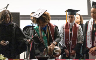 A woman in a graduation cap and gown pours water from a vase into a bowl on a table inside. Two other graduates watch, as a woman to the left holds a microphone.