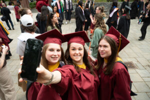 A group of three women in maroon graduation gowns and caps smile and take a selfie.