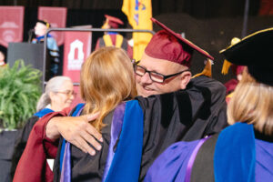A man in a maroon graduation cap and glasses hugs a woman in a black graduation gown.
