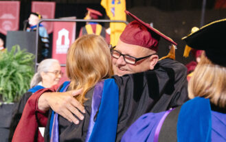 A man in a maroon graduation cap and glasses hugs a woman in a black graduation gown.