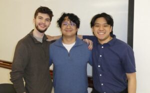 Three young men stand together inside a classroom and smile.