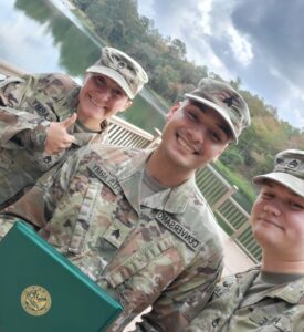 Three soldiers stand side by side smiling. The one in the middle is holding a green diploma cover. They are all in camouflage.