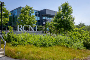 A silver statue of the letters RCNJ stands in front of green trees and a building.