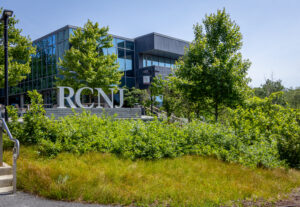 A silver statue of the letters RCNJ stands in front of green trees and a building.