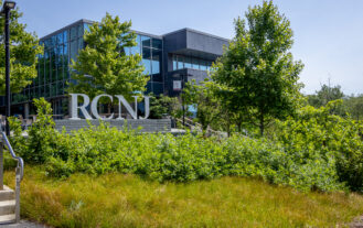 A silver statue of the letters RCNJ stands in front of green trees and a building.