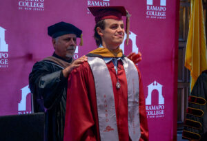 A young man in a maroon graduation gown and cap stands as a man in a black graduation gown and cap places a yellow hood around his shoulders.