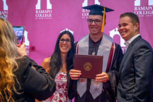 Three people stand in front of a maroon backdrop stating Ramapo College of New Jersey. The young man in the middle wears a graduation gown and cap and holds a diploma. He is flanked by a smiling woman and man. On the left is someone taking their photo on a phone.