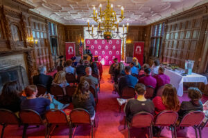A room with wooden walls and a large chandelier is filled with about 30 maroon chairs and people sitting on them. At the front of the room is a person in a graduation gown at a podium.