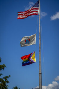 A flag poll holds three flags. On the top is the American Flag, middle is NJ State Flag, and on the bottom is the Pride Flag. The three flags fly against a blue sky.