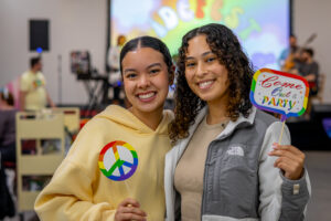 Two young women stand side by side smiling. One is holding a rainbow peace sign. The othe is holding a rainbow sign that reads "Come out & party"