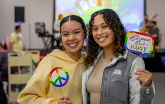 Two young women stand side by side smiling. One is holding a rainbow peace sign. The othe is holding a rainbow sign that reads "Come out & party"