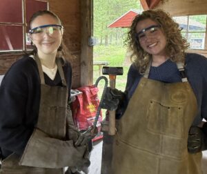 Two young women pose in a blacksmith shop. They have on brown aprons and eye protection.