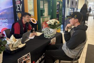 A person in a red shirt and black vest sits behind a table taking notes, as someone at the other side of the table talks to him. They are inside a supermarket.