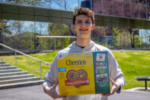A young man holds a Cheerios box in front of him as he stands outside and smiles. He is in front of a mirrored building.