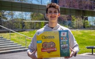 A young man holds a Cheerios box in front of him as he stands outside and smiles. He is in front of a mirrored building.