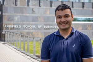 A young man stnands outside in front of a building and smiles.