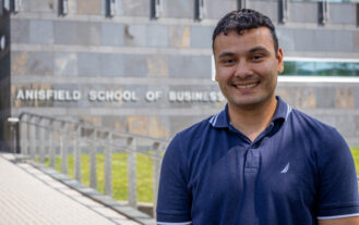 A young man stnands outside in front of a building and smiles.
