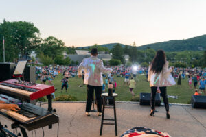 The image was taken from behind the band performing to a crowd seated & standing on the open green lawn. There are two keyboards in the left-hand corner and two performers centered in the image wearing white sequined jackets and black pants.