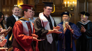 A pair of young men wearing graduation gowns and caps stand and clap. A few people around them stand, clap and look at them.