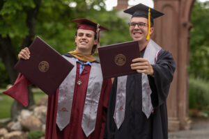 Two young men in graduation gowns and caps hold diplomas in front of them. They are standing outside.