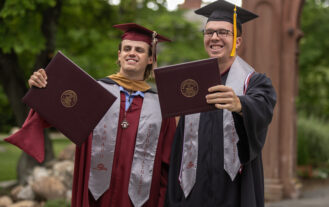 Two young men in graduation gowns and caps hold diplomas in front of them. They are standing outside.
