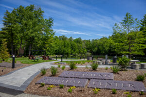 A pond is surrounded by trees, with a blue sky overhead. In front is a path of pavers, with grass and maroon chairs. A pair of large rectangular stones has the words Salameno Spiritual Center etched in them.