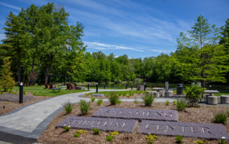 A pond is surrounded by trees, with a blue sky overhead. In front is a path of pavers, with grass and maroon chairs. A pair of large rectangular stones has the words Salameno Spiritual Center etched in them.