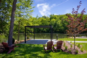 An open air pergola sits on a patio and next to a pond, with green trees around it. Maroon chairs are in the surrounding grass.