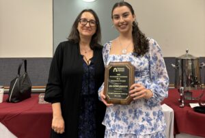 Two women stand next to eachother inside. The one on the right is holding an award.