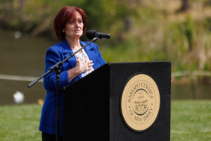 A woman stands at a podium outside in front of a pond. She has her hand on her heart.