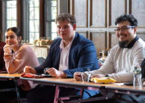 Three college students sits at a table and listen. They are inside, with windows behind them.