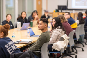 Students seated at a conference table for class, looking at the professor (who is off-screen).