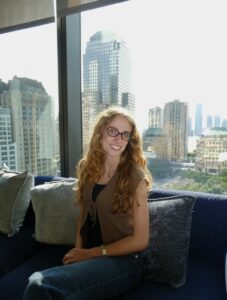 A woman sits in front of a large window with the New York City skyline behind her.