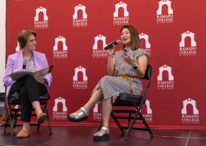 Two women sit on chairs on a stage in front of a maroon banner with Ramap College logos. One holds a microphone and speaks as the other looks at her and listens.