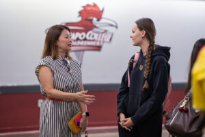 A woman speaks with a young woman. They are standing and are inside, in front of a Ramapo College logo on a wall.