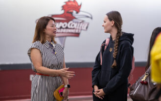 A woman speaks with a young woman. They are standing and are inside, in front of a Ramapo College logo on a wall.