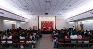 A crowd of people sit in chairs in front of an indoor stage. On stage are two women sitting in front of a maroon Ramapo College banner.