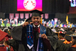 A student in cap and gown is smiling at the camera walking down the aisle at the 2026 commencement ceremony. The stage is in the background with the RCNJ logo on a large screen.