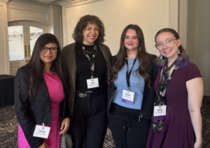 A group of four women in business clothes stand side by side. They are inside.
