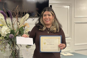 A woman stands inside a ballroom and holds a certificate in one hand and an envelope in the other. She is smiling.