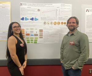 A woman and a man flank a research poster on a wall. They smile.