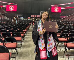 A woman with wearing a graduation cap and gown stands inside a stadium holding a maroon diploma and smiling.