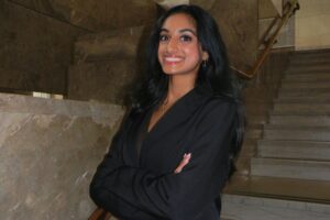 A woman wearing a black business suit stands in front of stairs, crosses her arms, and smiles.