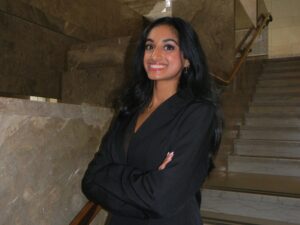 A woman wearing a black business suit stands in front of stairs, crosses her arms, and smiles.