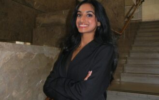 A woman wearing a black business suit stands in front of stairs, crosses her arms, and smiles.
